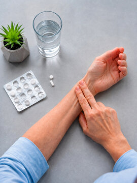 Woman checking pulse on her wrist. Senior person measuring heart rate with finger. Medical self care concept with pill and glass of water on table. Healthcare monitoring at home.