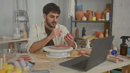 Fototapeta premium Man sculpts pink clay pot with hands on a spinning pottery wheel in studio, using loop tool and sponge while watching a laptop; concentration creativity.