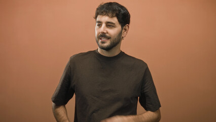 Fototapeta premium Man with beard crossing arms and slight smile in studio portrait with warm terracotta wall and dark tshirt; quiet confidence.