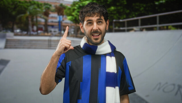 Young bearded man in football shirt and scarf points finger upward on street in front of skate ramp; pride camaraderie.