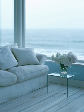 A serene coastal living space featuring a white linen sofa, glass side table with fresh flowers, and a panoramic view of the calm blue sea through large windows.