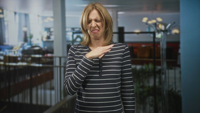 Woman covers nose with hand and recoils in a building atrium near railing and lamps while grimacing; disgust rejection.