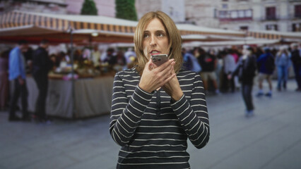 Fototapeta premium Woman holding smartphone to mouth speaking into speakerphone with expressive hand gesture among crowded outdoor market stalls; frustration urgency.