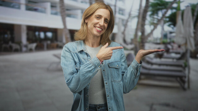 Woman in denim jacket points with open palm to poolside lounge chairs beside a hotel building, smiling and slightly turned to the side; inviting welcome.