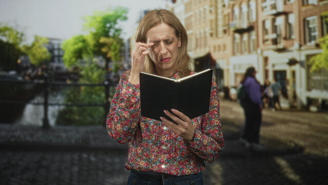 Woman holds a black book, finger rubbing near eye while reading on street by canal; concentration frustration.