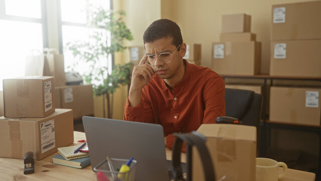 Man in a small business building clasps hands under chin over laptop amidst stacked boxes and a scanner on desk, wearing glasses and looking pensive; anxiety.