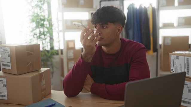 Man holding smartphone to ear among stacked shipping boxes and laptop in a warehouse building, checking orders on a laptop; concern responsibility.