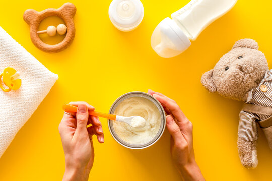 Make mixture for feed baby. Spoon with food in hand near toys and accessories on yellow background top view pattern