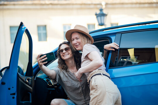 Two women taking a selfie beside a blue car during a road trip. Happy travel companions capturing memories with a smartphone on a sunny summer day