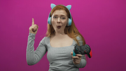 Redhead woman wearing blue headphones and a gray top holds a gamepad and points finger upward in a studio magenta backdrop  playful fun. © Krakenimages.com