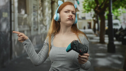 Redheaded woman holding a gamepad and wearing cat ear headphones points finger in a street  frustration defiance. © Krakenimages.com