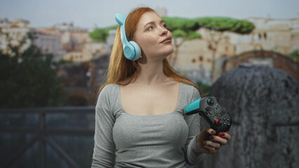Redheaded woman holding a gamepad and wearing blue headphones, head tilted and smiling in front of a building  playful. © Krakenimages.com