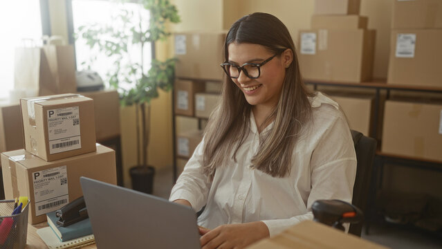 Woman typing on laptop among stacked shipping boxes and barcode scanner in office building; productive small business.