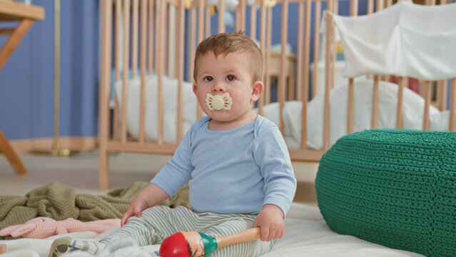 Toddler boy sitting with pacifier and holding maraca reaches hand toward crib in a building; innocent curiosity.