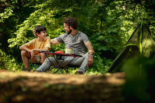Father and son enjoy a camping trip together in the woods with food and drinks on a table