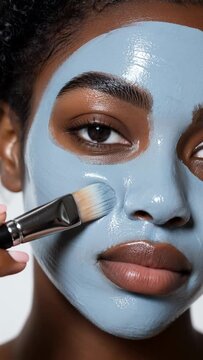 Close-up of a beautiful young Black woman applying a hydrating blue clay facial mask with a cosmetic brush for skin care and wellness.