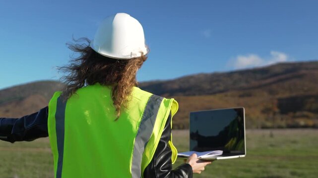 Engineer safety planning professional worker inspecting vast landscape site with hard hat and laptop computer on sunny day.