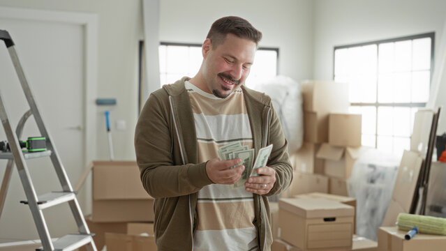 Man smiling while counting cash amid stacked moving boxes, ladder, and packing supplies inside a bright building room; savings relief.