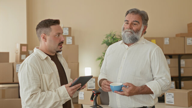 Two men hold a tablet and a coffee cup while one points with his hand at the screen amid stacked parcels in a building; focused teamwork.