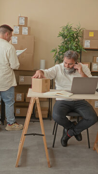 Man holding cardboard package with hand at small business building desk, talking on smartphone while checking laptop and inventory; focused teamwork.