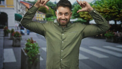 Obraz premium Man with beard using hands to frame face with thumb and forefinger on a pedestrian street, smiling at camera; spontaneous playfulness.