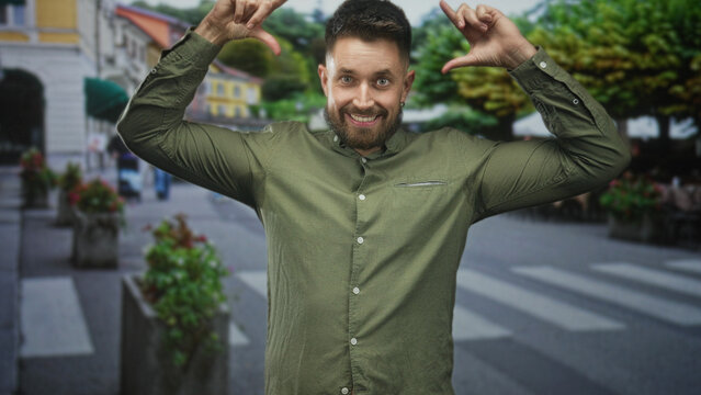 Man with beard using hands to frame face with thumb and forefinger on a pedestrian street, smiling at camera; spontaneous playfulness.