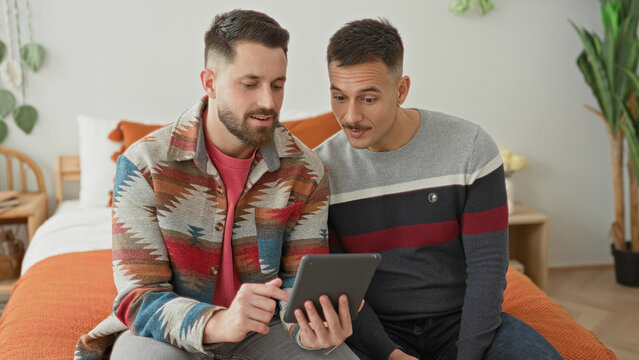 Two men on bed, one points finger to tablet while both lean in viewing the screen in bedroom; comfort connection.