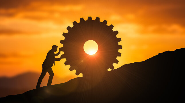 Determined businessman silhouette manually pushing giant heavy gear wheel up steep mountain incline against vivid orange sunset representing intense physical struggle and progress