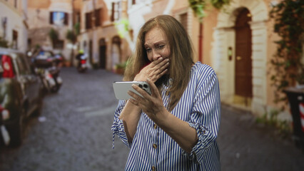 Fototapeta premium Senior woman holding smartphone with hand to mouth in amazed smile on a cobblestone street; surprise memory.