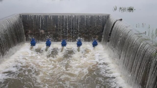 A concrete weir structure controls water flow in a rural river. The storage system features blue valves releasing stream current during irrigation supply management for agriculture.