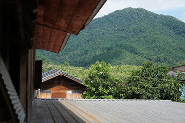Obraz premium elevated perspective framed by traditional wooden house eaves, looking out across corrugated metal rooftops toward a massive, vibrant green mountain. The composition highlights peaceful countryside li