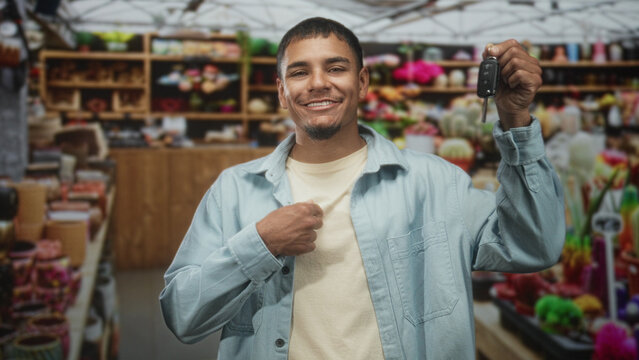 Man holds car keys aloft and presses chest with other hand in a retail building, smiling and asserting ownership; confidence success.