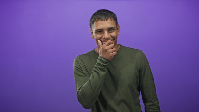 Man with hand covering mouth in a shy smile, wearing green shirt in studio with purple backdrop and soft lighting; playful warmth.