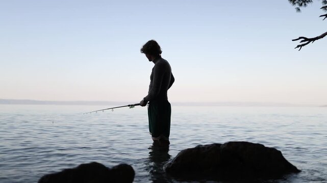 Young man stands knee deep in tranquil sea water holding a fishing pole and watching the float while patiently waiting for a bite during serene evening fishing near shore