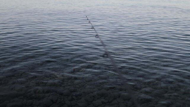 Fishing rod points toward a small float resting on clear sea water while the angler waits for a bite above a rocky seabed during a calm peaceful evening fishing session