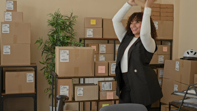 Woman reaches up with hands to grab a small cardboard box from stacked parcels on shelving in a building, smiling and organizing packages; entrepreneurial joy.