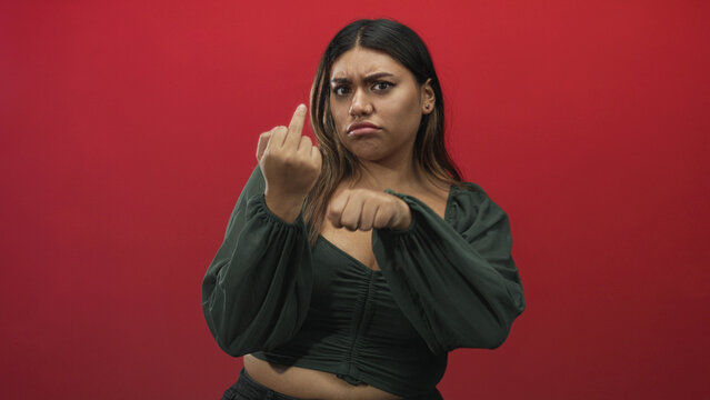 Woman plus size in green blouse makes a fist and shows middle finger in studio with red backdrop and tense expression; defiance anger confrontation.
