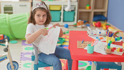 Naklejka premium Little girl cutting paper with orange scissors at a red table in building surrounded by toys and art supplies; creative concentration play.