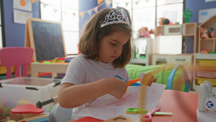 Girl cutting paper with child scissors at a colorful preschool classroom table, hands holding scissors and drawing sheet, wearing a plastic tiara and white shirt  concentration. © Krakenimages.com