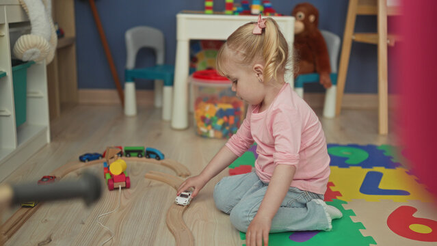 Little girl kneeling on foam mat pushes a white toy car along a wooden train track in a playroom building; playful curiosity.
