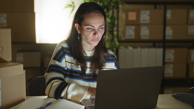 Woman points at notebook and types on laptop amid stacked shipping boxes and parcels in a building; late night focus.