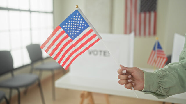 Woman holding small american flag with visible hand at a pollingstation voting booth; pride civic duty.