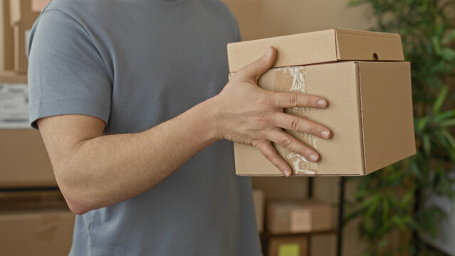 Man holding stacked cardboard boxes with visible hand and fingers while carrying packages in building storage area with shelves and plant visible; delivery service diligence.