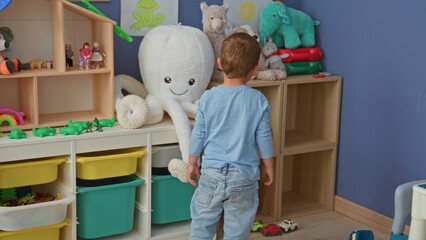 Fototapeta premium Boy picking up a yellow bucket next to a large plush octopus on shelves in a building playroom; curiosity play.