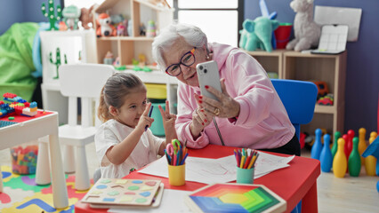 Fototapeta premium Woman holding smartphone taking selfie with little girl at colorful preschool classroom table showing drawings and colored pencils and toys on shelves; generational bonding joy.