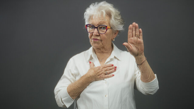 Elderly woman with glasses places hand on chest and raises palm in oath gesture in studio; honor sincerity.