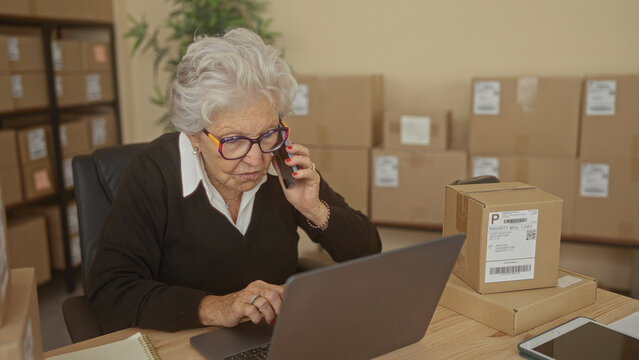 Elderly woman holds phone to ear and types on laptop amid stacked shipping boxes and pallet shelves in a building; determination entrepreneurship.