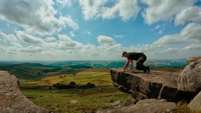 Hiker climbing onto dramatic rock ledge above wide valley landscape. Outdoor adventure on rugged granite outcrop with panoramic countryside and expansive mountain scenery. Active travel, hiking