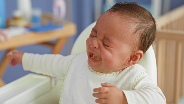 Hispanic baby boy in highchair being spoon fed, food on mouth and reaching with scrunched face, crying gesture in a building; frustration refusal.