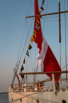 Flags and sails on a ship during sunset over the water. A ship displays various flags and sails against a sunset sky over the water, with its rigging visible.